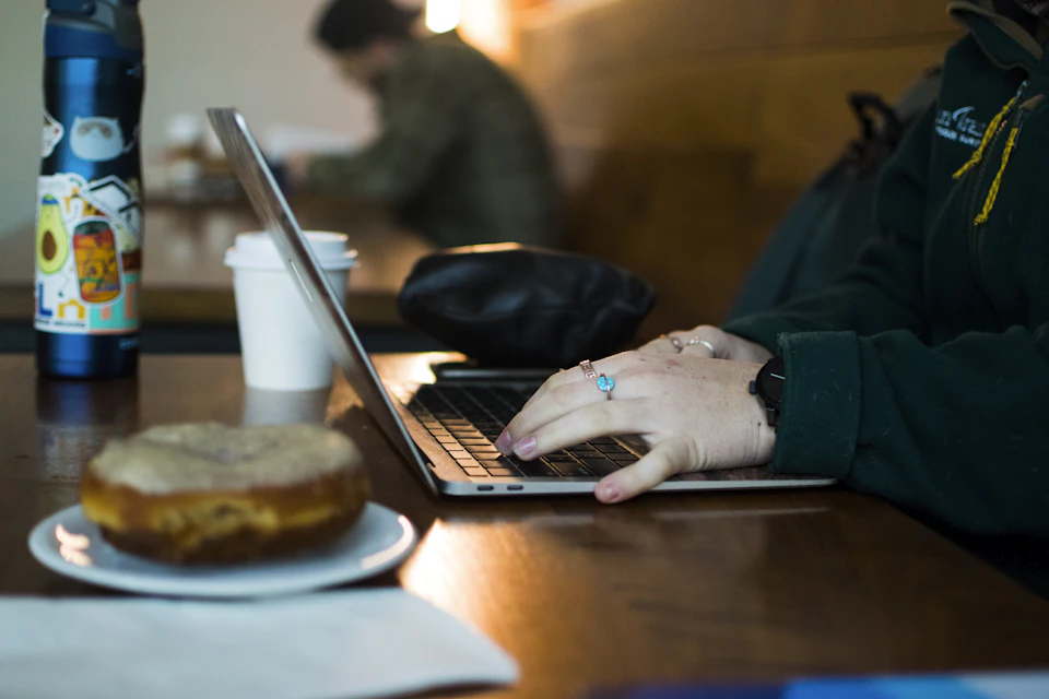 Persona che usa il portatile al tavolo di un bar, con ciambella e caffè